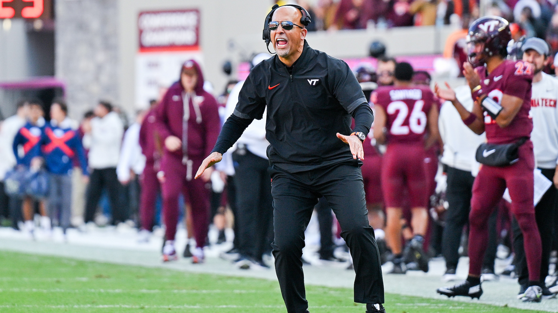 A football coach in black attire and sunglasses is actively directing play on the field during a game.