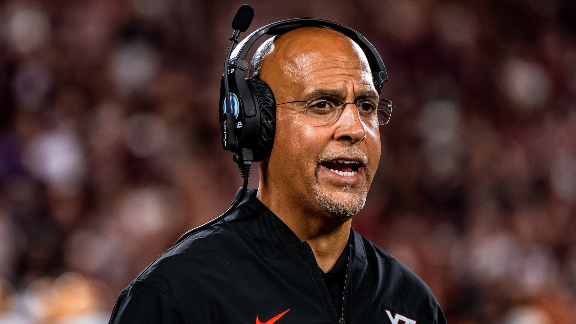 A coach wearing a headset and a black jacket with a Nike logo and VT emblem is speaking during a game, with a blurred crowd in the background.