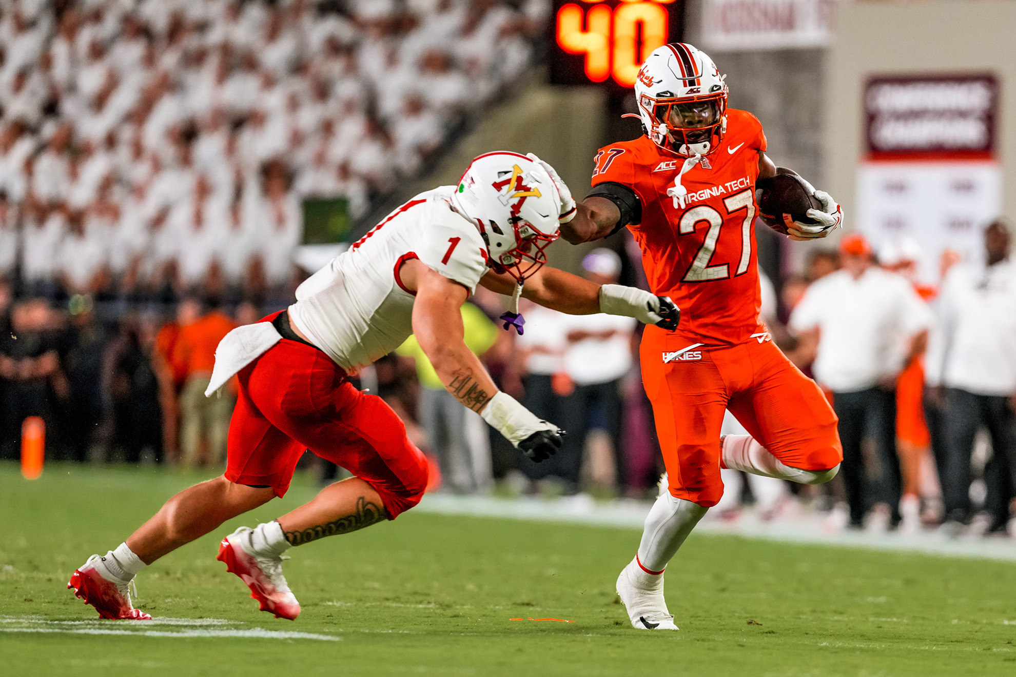 A football player in an orange uniform with the number 27 runs with the ball while being tackled by an opponent in a white and red uniform.