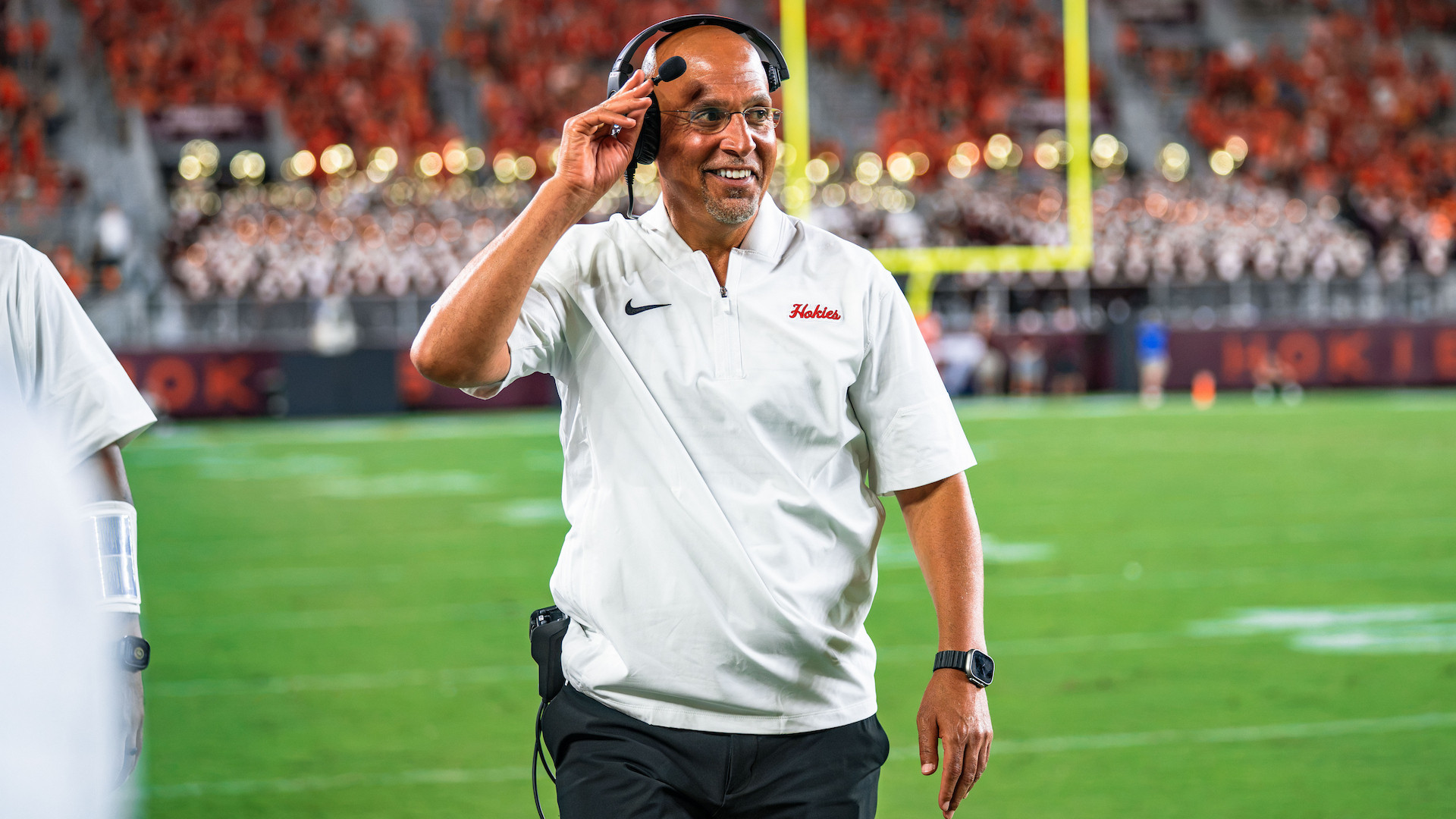 A coach in a white polo shirt and headset smiles while adjusting his headset on a football field during a game.