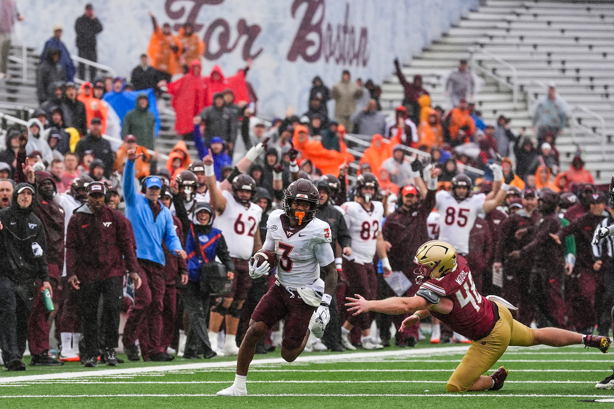 A football player in a white jersey with the number 3 runs with the ball as a defender in a maroon jersey dives to tackle him on a snowy field.