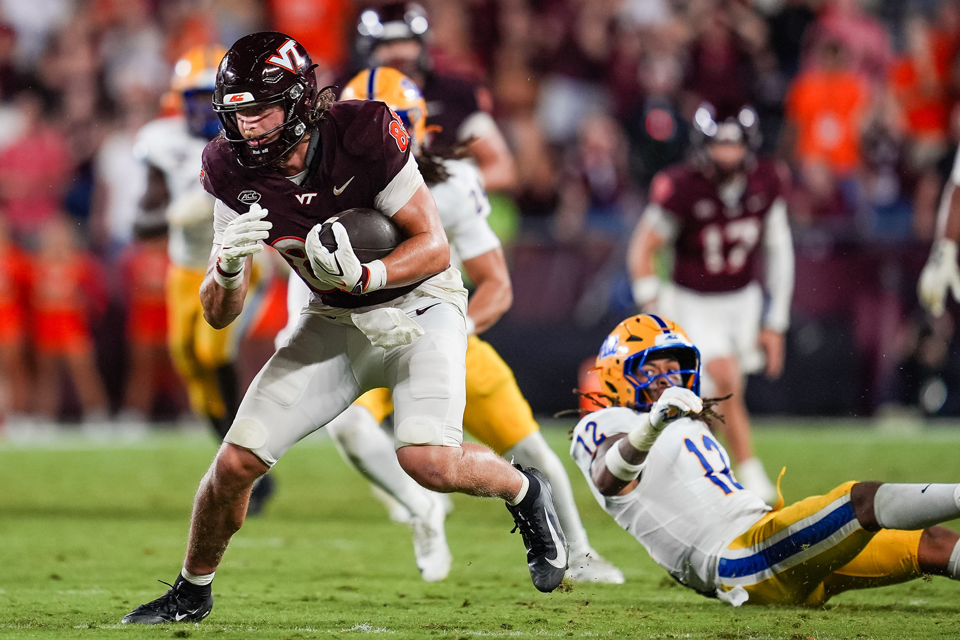 A football player in a maroon jersey runs with the ball while being pursued by an opponent in a white and yellow uniform.